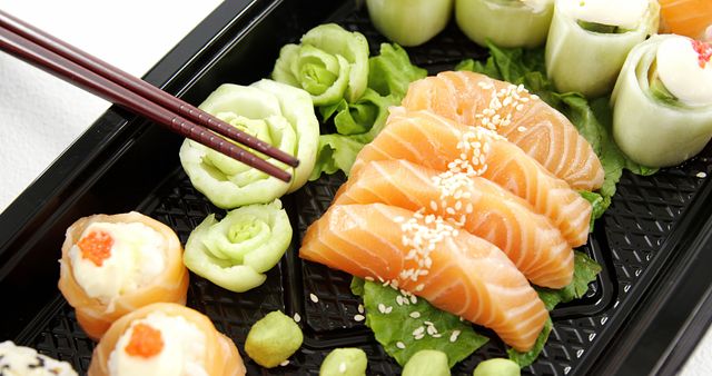 Chopsticks are poised to pick up a slice of salmon sashimi from a bento box filled with an assortment of sushi and rolls, garnished with green lettuce and wasabi. A delicious Japanese cuisine selection, perfect for a fresh and healthy meal option.