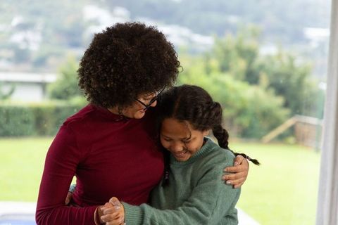 African american mother hugging daughter near sliding glass door with backyard view