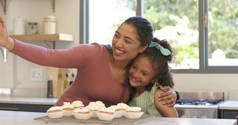 Diverse Mother and Daughter Taking Selfie and Smiling with Cupcakes in Sunny Kitchen