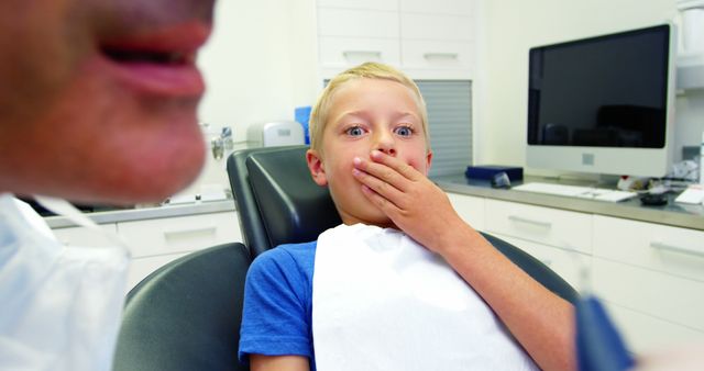 Young boy covering mouth with hand while sitting in dental chair, showing visible anxiety as dentist tends to him. Dentist, partially visible, bringing instrument close to patient. Modern dental clinic equipment and background create professional atmosphere. Useful for articles on pediatric dentistry, dental anxiety in children, healthcare settings, and dental clinic advertisements. Can be utilized for topics addressing emotional responses to medical environments or healthcare services for children.