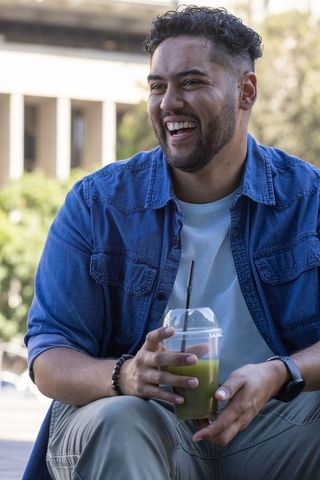 African american man laughing holding green juice on urban plaza bench in denim shirt