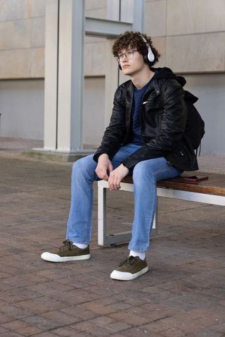 Thoughtful teen male sitting on bench listening to music with headphones in urban plaza
