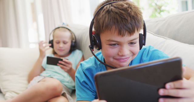 Children sitting on a sofa, using tablets and smartphones while wearing headphones. Boy in blue shirt focused on tablet in foreground, girl reclining and using smartphone in background. Ideal for themes related to technology, digital lifestyle, family activities, and modern entertainment. Perfect for articles, blogs, or advertisements about parenting, digital childhood, educational apps, or home-based relaxation.