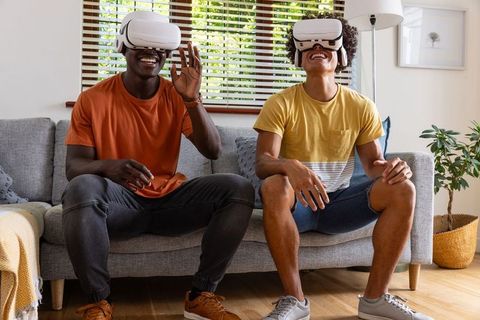 Diverse male friends enjoying virtual reality headsets on sofa, laughing and interacting