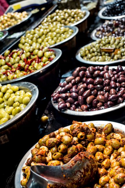 This captures a vibrant display of various marinated olives in stainless steel bowls on a market stall. Ideal for use in content related to Mediterranean cuisine, gourmet food, healthy eating, market scenes, and culinary displays.