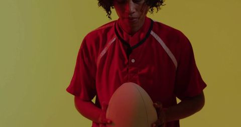 Adult woman holding oval ball and gazing down in red jersey on yellow backdrop