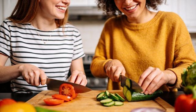Two women slicing fresh tomatoes and cucumbers together on wooden cutting board for meal prep