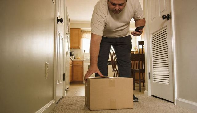Man bending to retrieve package in hallway while checking smartphone for delivery status