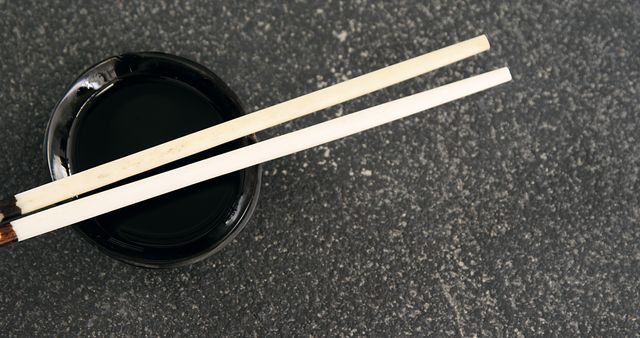 Chopsticks resting on soy sauce bowl over grey stone surface provide an elegant minimalist presentation. Ideal for use in content related to Japanese or Asian cuisine, traditional dining, food blogs, and culinary arts. Useful as a visual element in restaurant menus, recipe illustrations, and food-related advertisements.