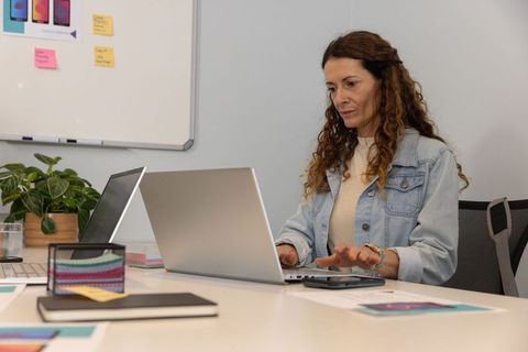 Mature woman in denim jacket typing on laptop and reviewing app mockups in modern office