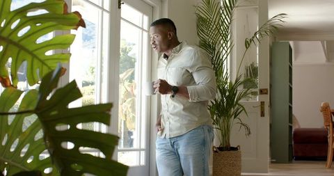 Sunlit african american man standing by glass doors holding coffee near indoor plants