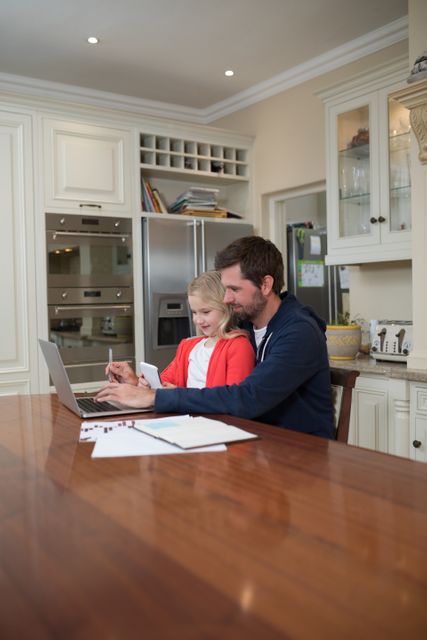 Father and daughter using a laptop together in a modern kitchen. Ideal for themes related to family bonding, technology in education, remote work, and modern family lifestyles. Can be used in articles, advertisements, or websites focusing on parenting, education, or home technology.