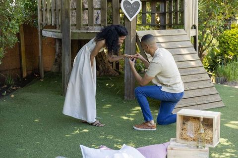 Kneeling man proposing ring to smiling woman in white dress, sunlit backyard engagement