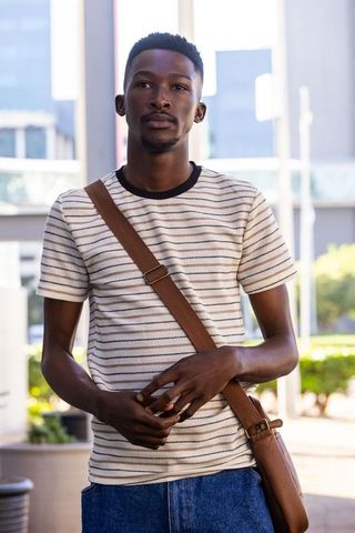 Young african american man standing in urban plaza wearing striped shirt and crossbody bag