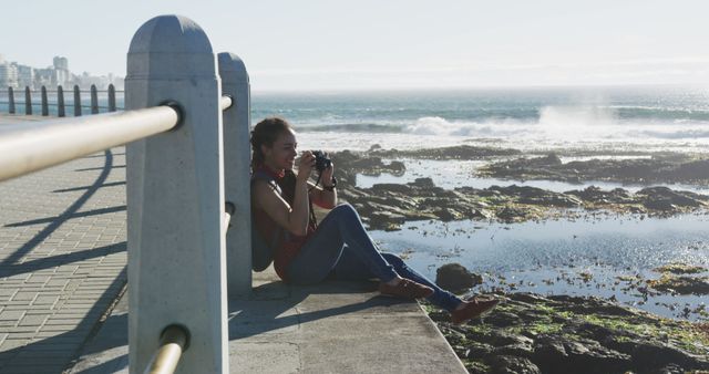 Woman sitting by the seaside with a camera, appreciating the coastal views. Can be used for travel blogs, adventure content, lifestyle advertisements, or articles promoting outdoor activities.