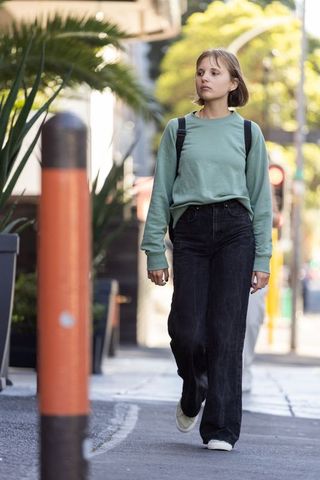 Young woman walking sidewalk wearing mint sweatshirt and black jeans carrying backpack