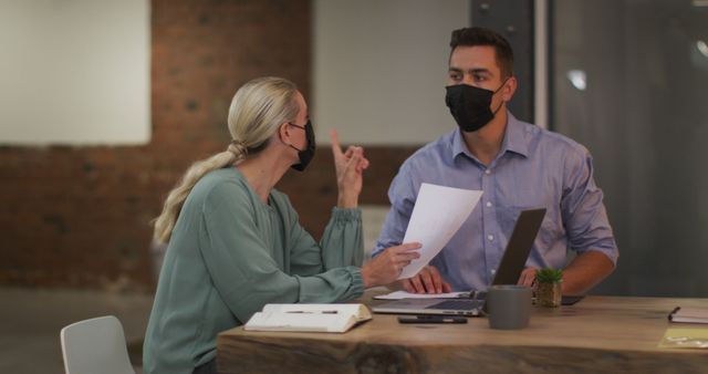 Three colleagues are seen in an office environment wearing masks, engaging in a work discussion at a table. This conveys a sense of pandemic precautions in a professional setting, highlighting collaboration and safety measures. Suitable for content focused on workplace safety, pandemic, teamwork, office collaboration, and business operations during COVID-19.