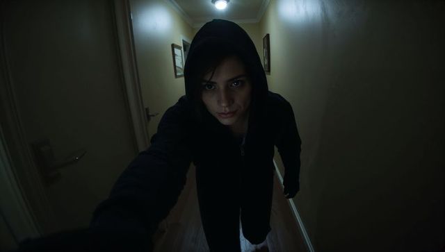 Hooded woman leaning toward camera in dim hallway reaching hand dramatic low-light portrait