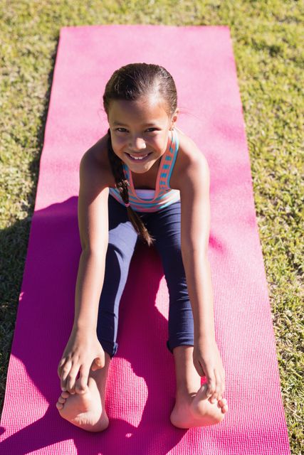 Young girl with braided hair practicing yoga on a pink mat in a park, smiling and stretching forward. Ideal for promoting children's fitness, outdoor activities, healthy lifestyle, and yoga classes for kids.