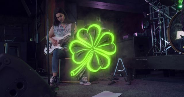 A young female musician is playing a bass guitar on a dimly lit stage while a glowing neon clover is displayed prominently beside her. This could be used to promote live music events, band promotions, youthful music culture, creative performances, or St. Patrick's Day themed concerts.