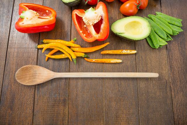 Colorful assortment of fresh vegetables including red peppers, avocado, cherry tomatoes, chilies, and snow peas arranged on a wooden table with a wooden spoon. Perfect for use in recipes, healthy eating blogs, cooking tutorials, or kitchen-related websites.