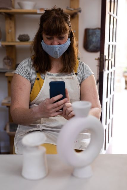 Caucasian female potter wearing face mask using smartphone in pottery studio. Ideal for content related to health and hygiene practices in creative workspaces during the COVID-19 pandemic, artisan crafts, and the importance of safety measures in art studios.