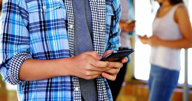 Teenager wearing plaid shirt focuses on smartphone while texting indoors. Out-of-focus individuals in background, suggesting a casual, social environment. Ideal for use in topics related to youth and technology, social interaction, mobile communication, and modern lifestyle.