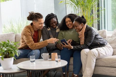 Cozy Friends Sharing Smartphone on Sofa in Sunlit Modern Living Room with Lush Plants