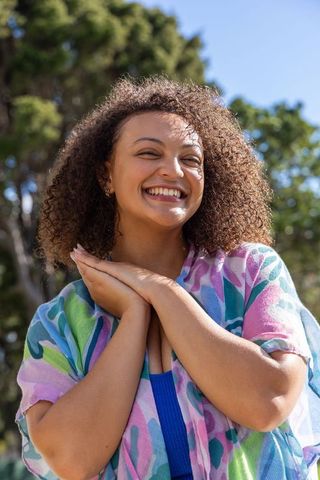 Joyful african american woman smiling in sunlit park wearing colorful patterned overshirt