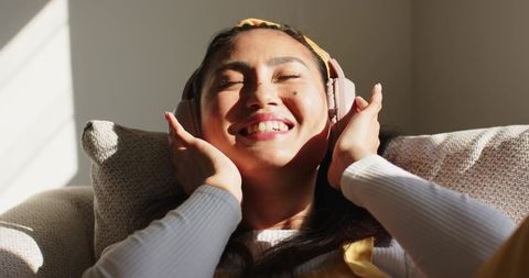 Sunlit asian woman smiling and listening to music with pink headphones on sofa at home