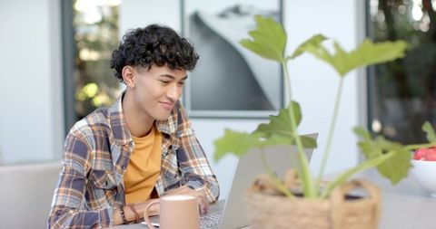 Young man working remotely on laptop at sunlit home table with plant and coffee
