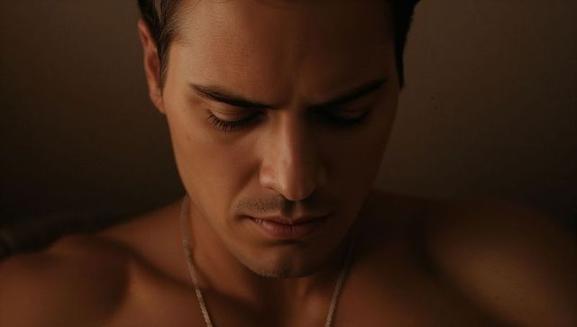 Shirtless hispanic man lowering head and closing eyes with silver chain in moody studio portrait