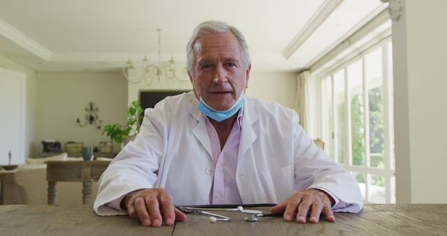 Senior male dentist sitting at table wearing white coat and mask, demonstrating dental tools indoors. Ideal for use in health-related articles, dental clinic advertisements, or portraying experienced medical professionals.