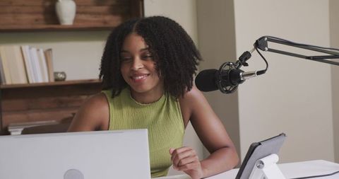 African american woman podcasting and smiling while working from home studio with laptop