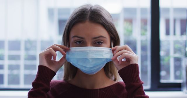 Young woman wearing a surgical mask standing indoors, lifting it to cover her face. Conceptually useful for topics related to health, safety, pandemic precautions, and protective measures. Ideal for healthcare, medical-themed posts, public health campaigns, and articles about virus prevention.