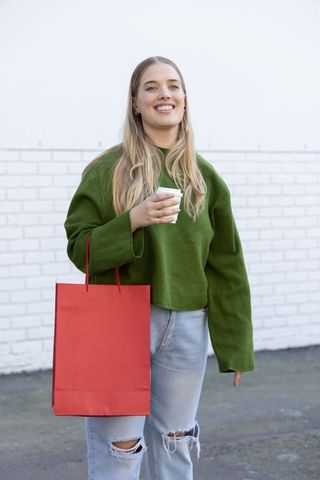 Young woman holding coffee and red shopping bag wearing green sweater and ripped jeans