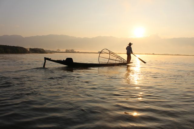 Silhouette of fisherman standing on a boat while paddling across a calm lake at sunrise. The scene is perfectly backlit by the setting sun, creating a serene and tranquil atmosphere with reflections on the water. This image is ideal for use in nature-themed publications, travel promotions, and materials emphasizing tranquility and peaceful outdoors. It captures traditional ways of life and can be used to convey themes of simplicity and harmony with nature.