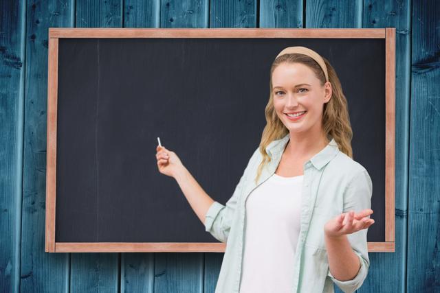 Blond woman standing in front of a blank blackboard, smiling, and pointing with chalk in hand. Can be used for educational content, training manuals, school promotions or advertisements, online teaching resources, or creative presentations.