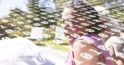 Laughing mature woman enjoying sunlit outdoor gathering with binary code digital overlay
