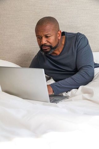 Mature african american man working on laptop in bed, focused remote work lifestyle