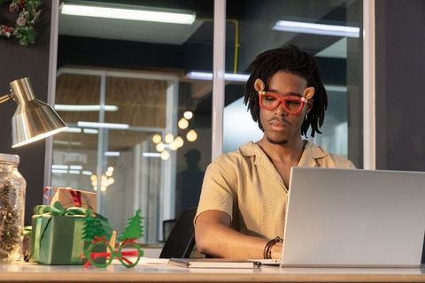 African american man working at office desk wearing festive novelty glasses using laptop