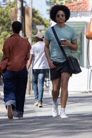Young man walking with coffee and crossbody bag wearing denim skirt and sunglasses casual street sty