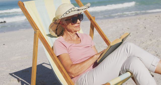 Elderly woman relaxing on a beach recliner while reading a book. She is wearing a wide-brimmed hat and sunglasses, enjoying a sunny day by the ocean. This scene can be used for materials related to leisure, retirement, vacation, healthy aging, and beach-related themes.