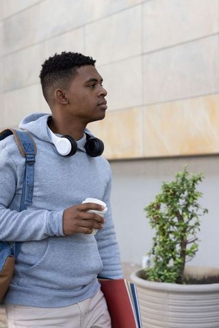 African american student holding coffee and notebooks wearing headphones on campus walk