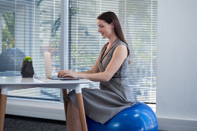 Female executive sitting on exercise ball while using laptop at desk in office