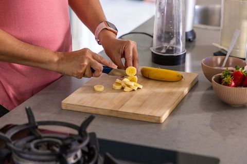 Woman slicing banana on wooden cutting board for smoothie preparation in modern kitchen