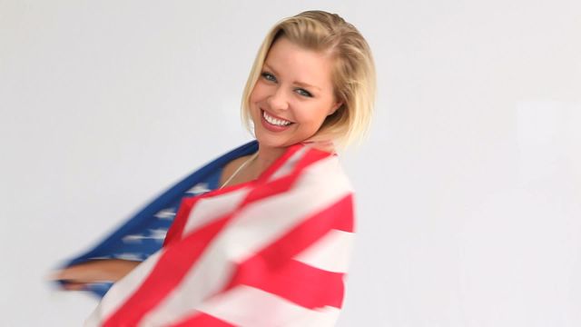 Blonde woman joyfully smiling while holding American flag. Ideal for content related to patriotism, national celebrations like Independence Day, USA-themed marketing, and promotional materials focusing on American pride and unity. Background is neutral, making it easy to use in various design contexts.