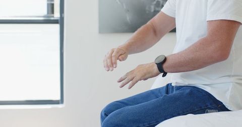 Mature man sitting on edge of bed wearing smartwatch in bright minimalist bedroom