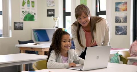 Female Teacher Assisting Smiling Student Using Laptop in Classroom