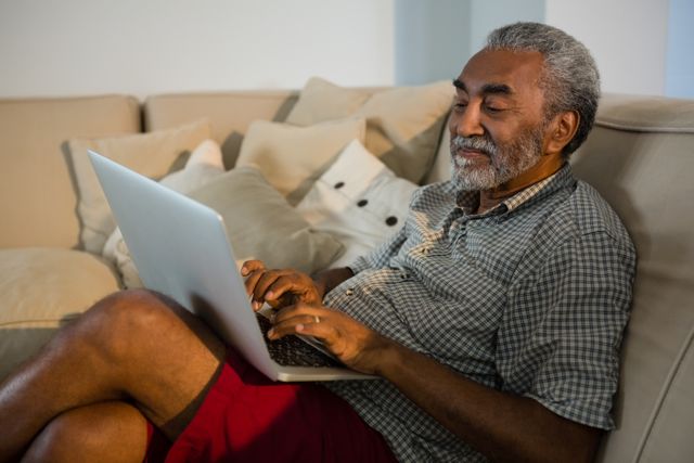 Senior man sitting on a sofa using a laptop in a comfortable living room. Ideal for content related to technology use among seniors, home lifestyle, leisure activities, and internet usage. Can be used in articles, blogs, or advertisements focusing on elderly people engaging with modern technology.